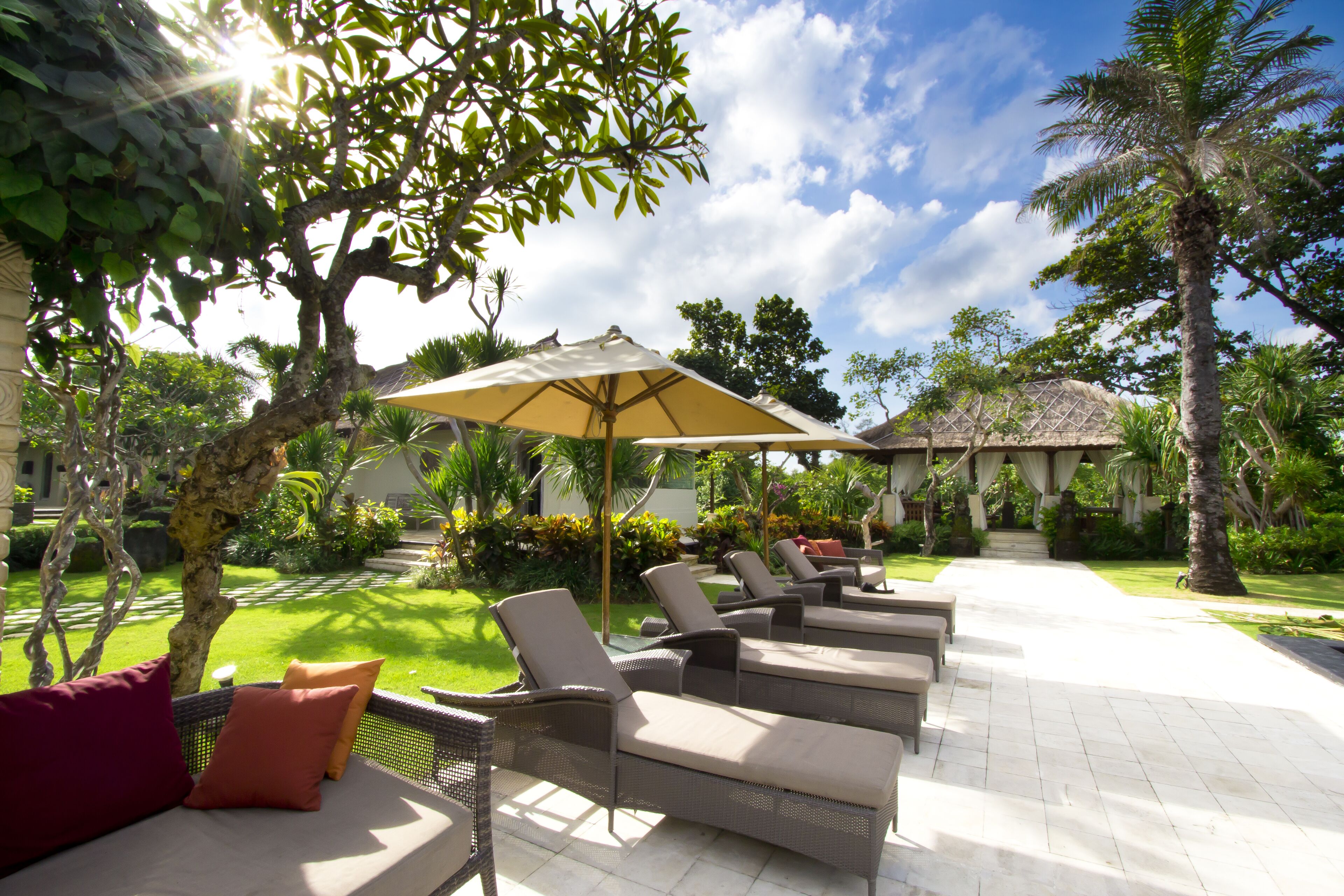 A sunlit outdoor seating area with lounge chairs and a large umbrella, surrounded by lush greenery and a clear sky.