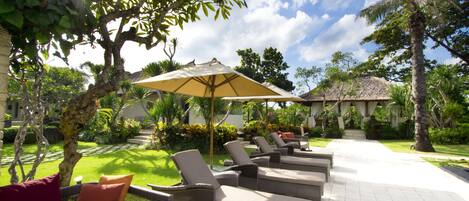 A sunlit outdoor seating area with lounge chairs and a large umbrella, surrounded by lush greenery and a clear sky.