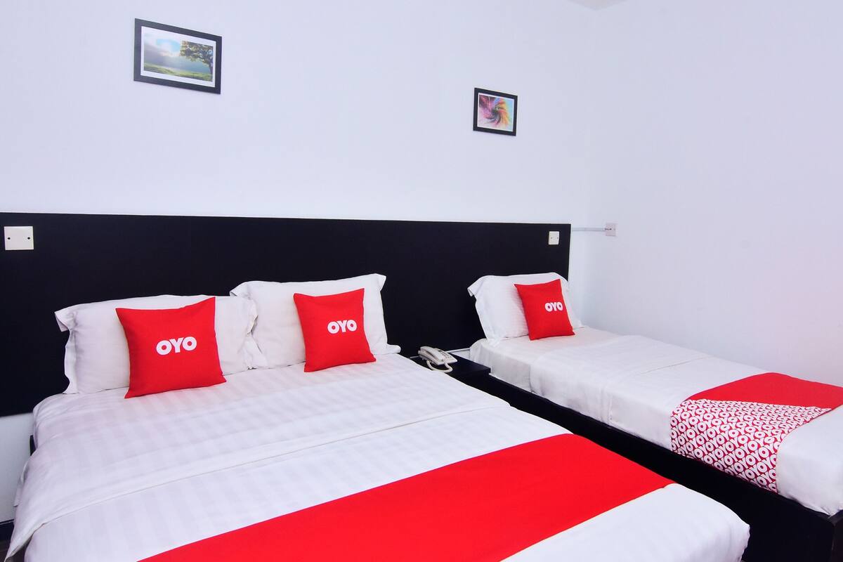 Habitación de hotel con dos camas, cada una con ropa de cama blanca y almohadas rojas con el logotipo 'OYO'. Hay dos cuadros en la pared, uno que representa un paisaje y otro un diseño abstracto y colorido.
