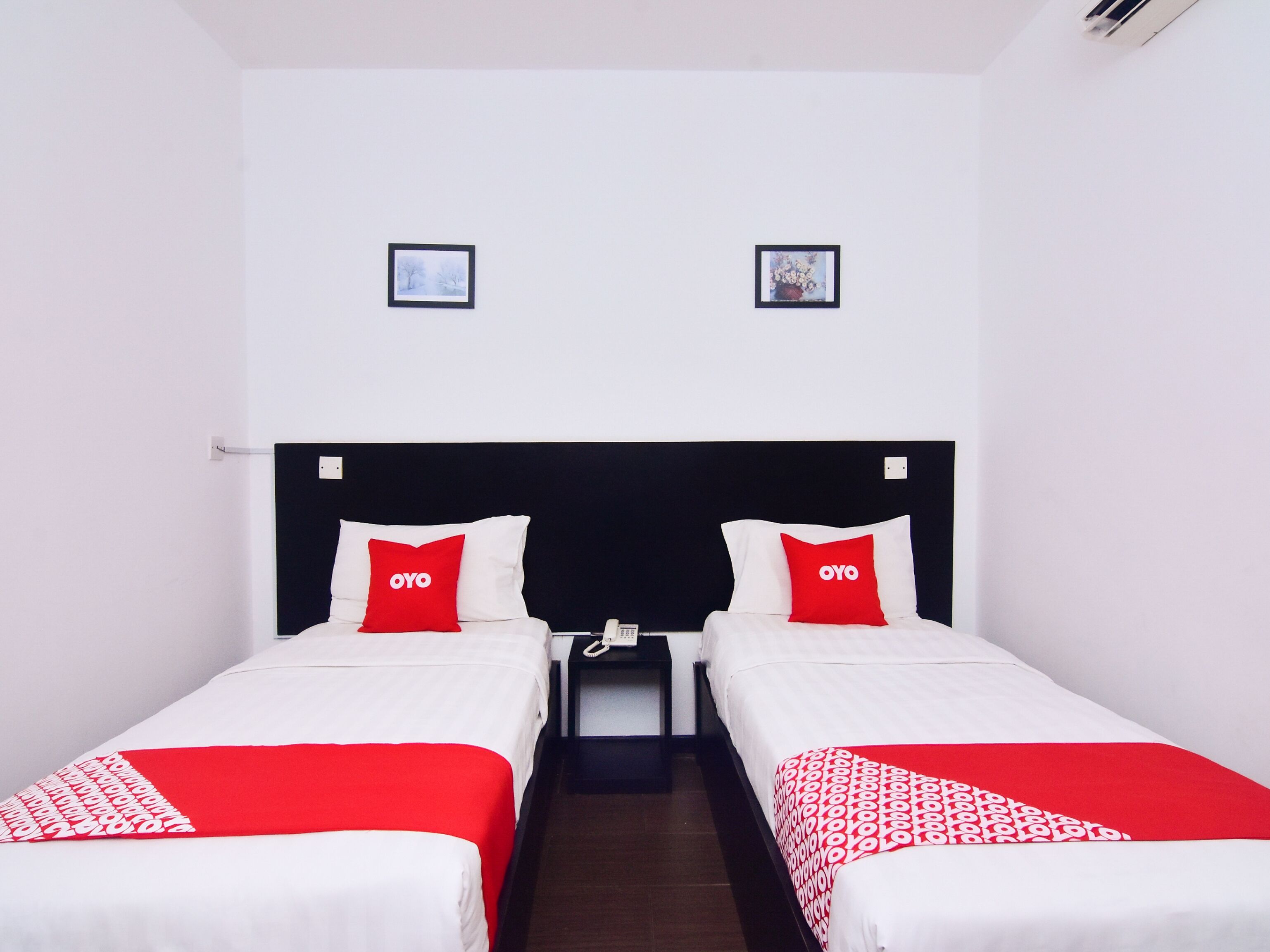 A hotel room with two beds, each with a red OYO pillow, a bedside table with a phone, and two framed pictures on the wall.