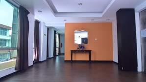 A hotel corridor with a wooden floor, large windows with curtains, an orange accent wall, and a small table with a vase.