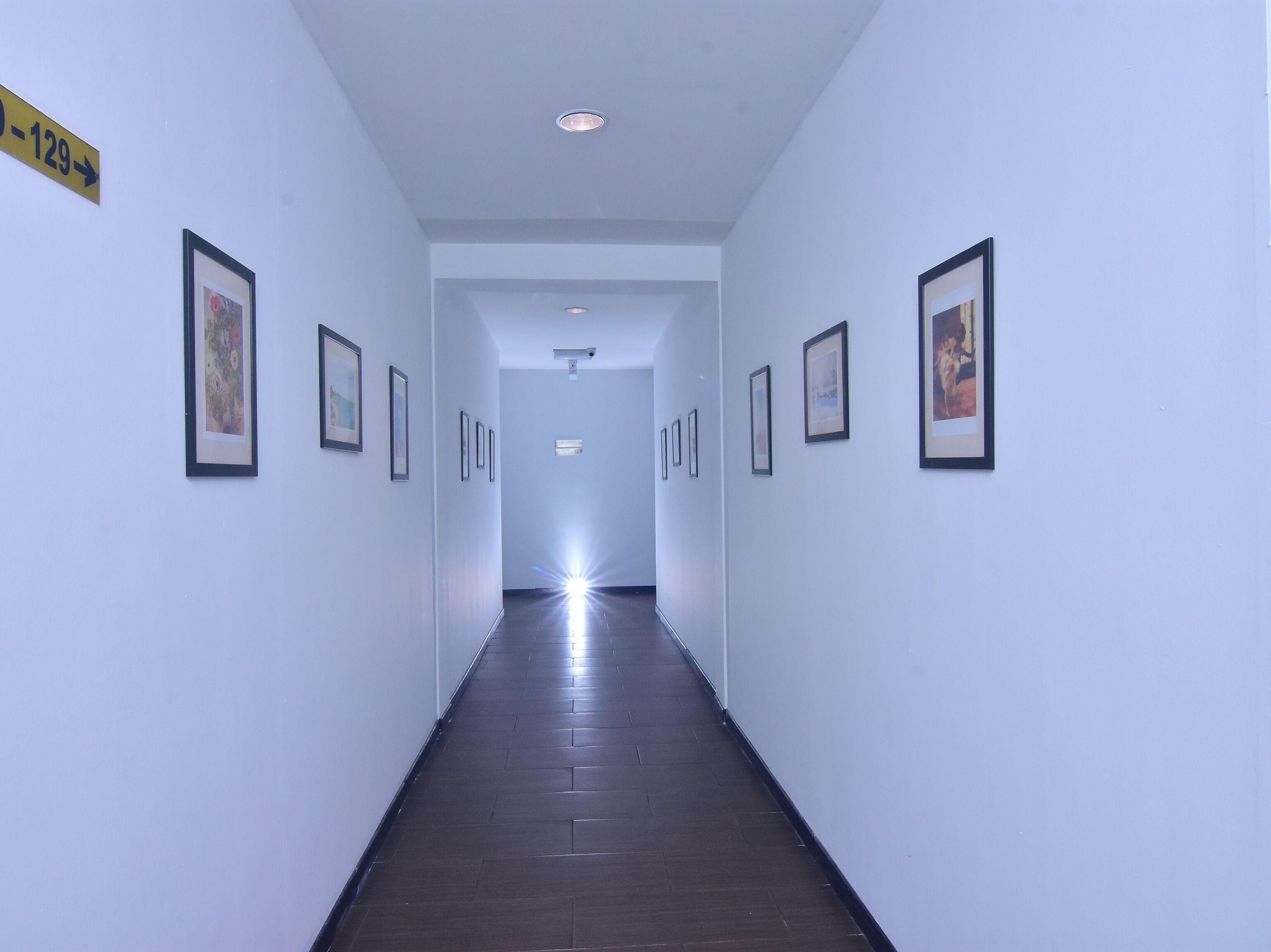A hallway with framed pictures on the walls, a sign indicating room number 129, and a ceiling light.