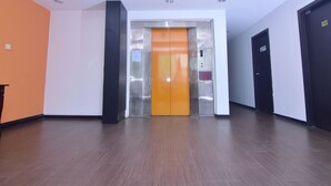A modern elevator with a metallic finish and a bright orange interior, situated in a hallway with wooden flooring and white walls.