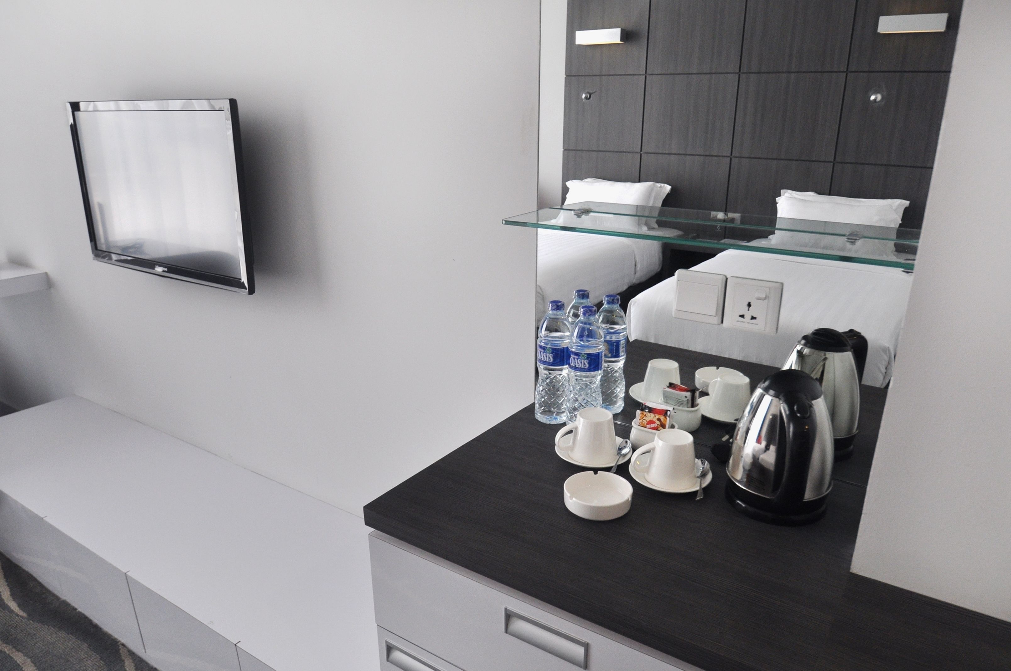 Una habitación de hotel moderna con una mesa de centro de vidrio, un televisor de pantalla plana en la pared, una cama con ropa blanca y una cafetera sobre la mesa.