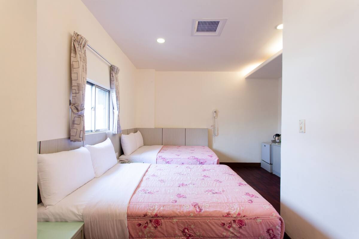 A hotel room with two beds, a window with curtains, a small kitchenette, and a ceiling vent.