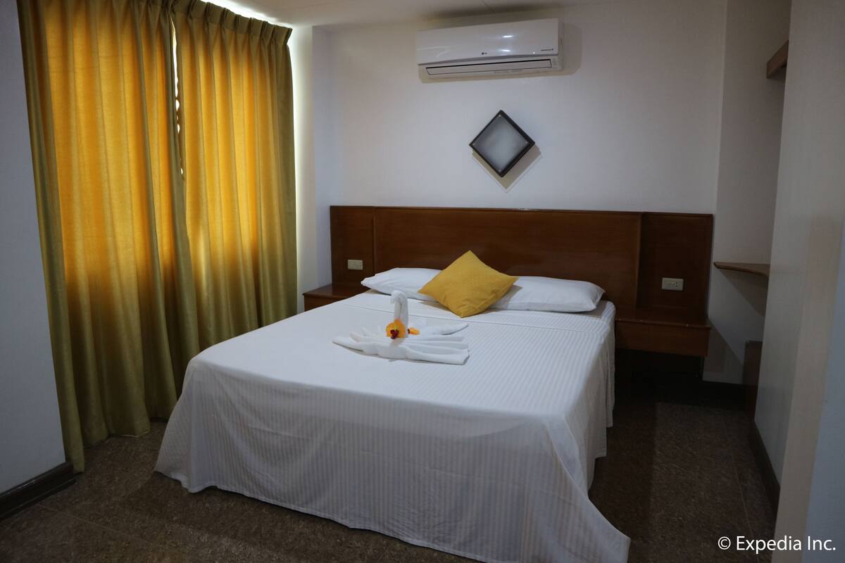 A hotel room with a neatly made bed, a single yellow pillow, and a swan-shaped towel arrangement.