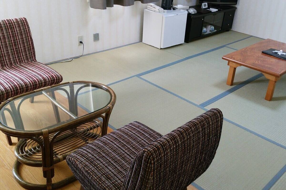 A living room with tatami flooring, a glass table, two chairs, a television, and a small white cabinet.