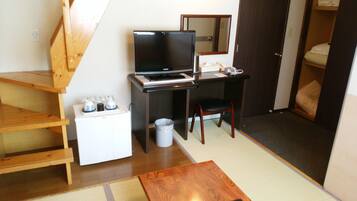 A compact room with a wooden desk, a television, a small refrigerator, a chair, and a bed in the corner.