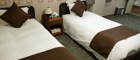 Habitación de hotel con dos camas individuales, cada una con un cojín marrón decorativo y una colcha blanca. Entre las camas, hay una mesita de noche de madera con una lámpara y un teléfono. El piso está alfombrado y hay una ventana con cortinas.