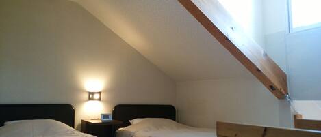 A bedroom with two beds, a nightstand with a lamp, and a sloped ceiling with exposed wooden beams.