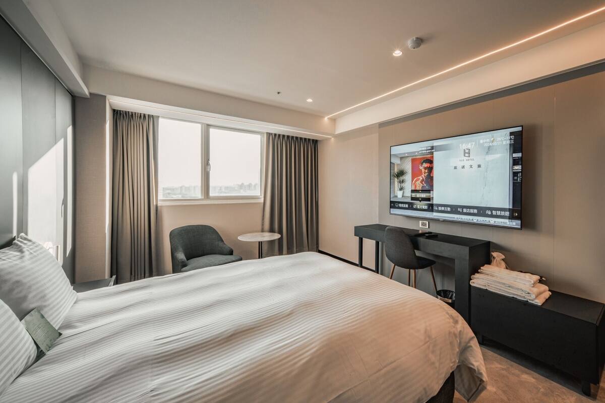 A modern hotel room with a large bed, a desk, a chair, a TV, and a view of the city.