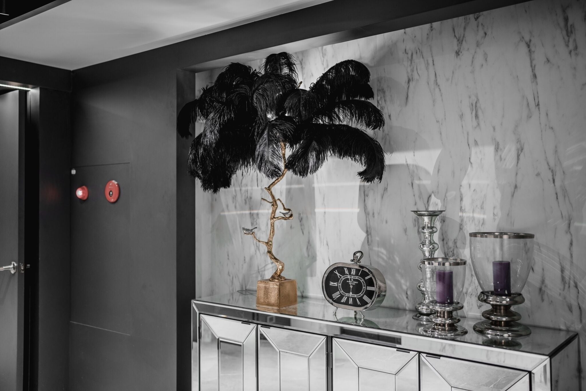 Un baño moderno con pared de mármol, una planta con plumas negras decorativas, un reloj y dos portavelas.