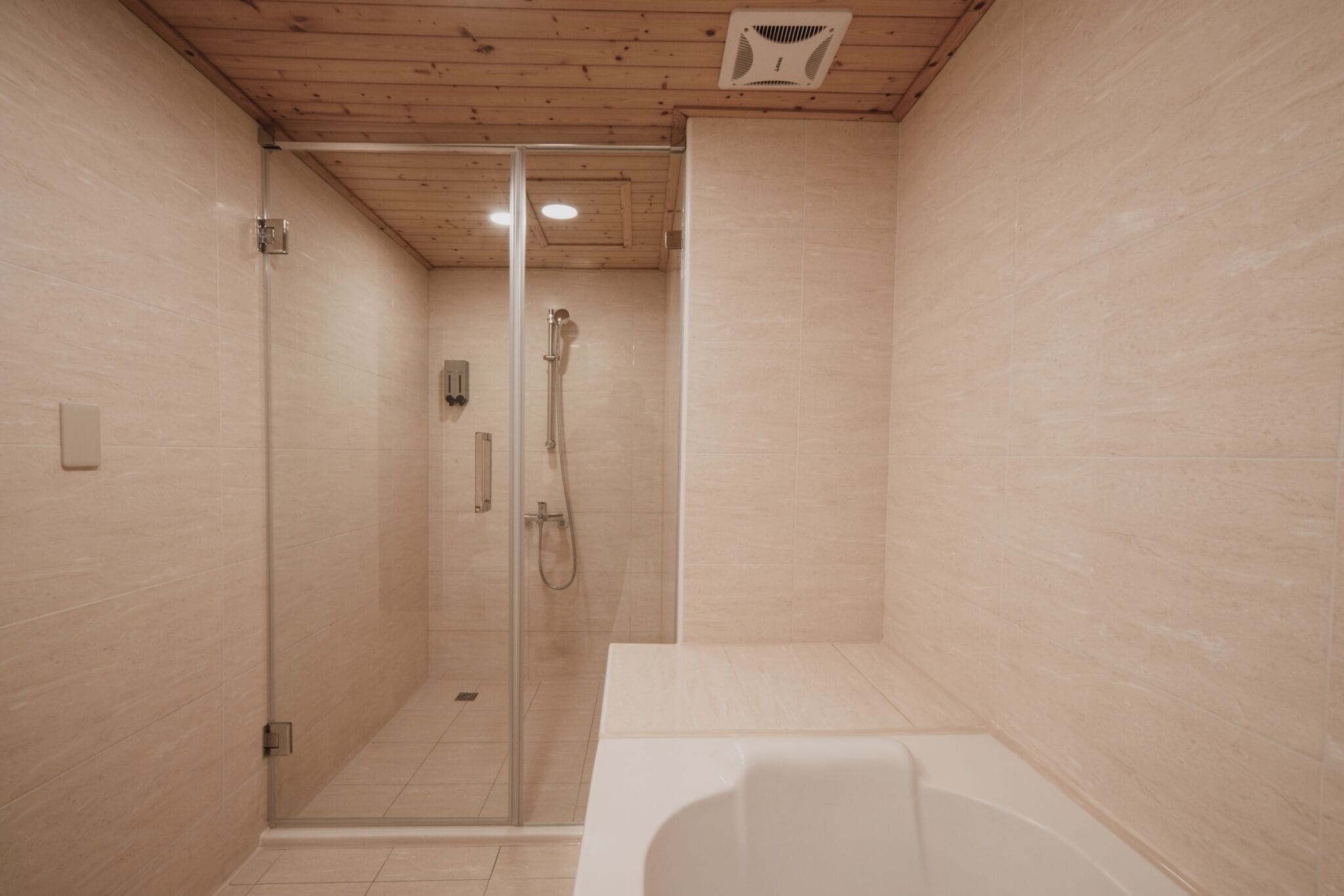 A modern bathroom with a glass-enclosed shower, a white bathtub, and beige tiled walls.