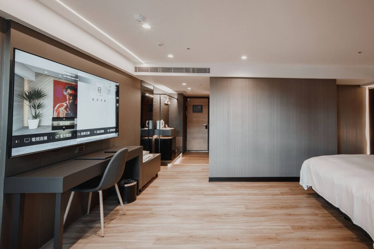 A modern hotel room with a large flat-screen TV, a desk, a chair, a bed, and a bathroom with a mirror and sink.