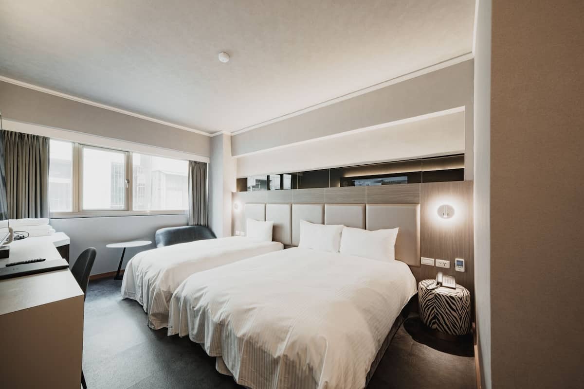 A hotel room with two beds, a desk, a chair, a small table, and a window with curtains.