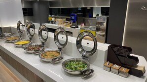 Un bufé con diversos alimentos en recipientes metálicos, una máquina de café y una caja de pañuelos.
