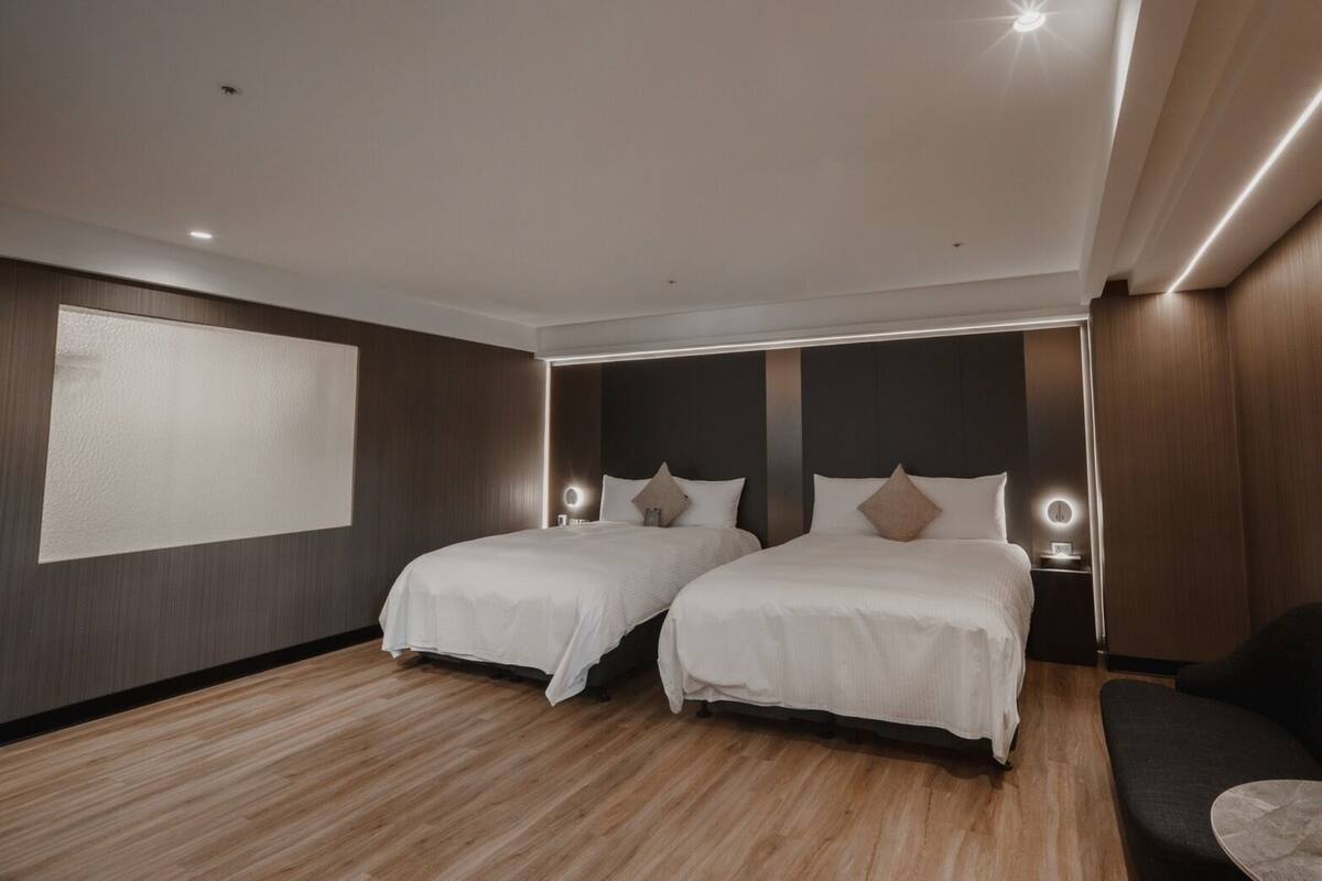 A modern hotel room with two beds, a large window, and a wooden floor.