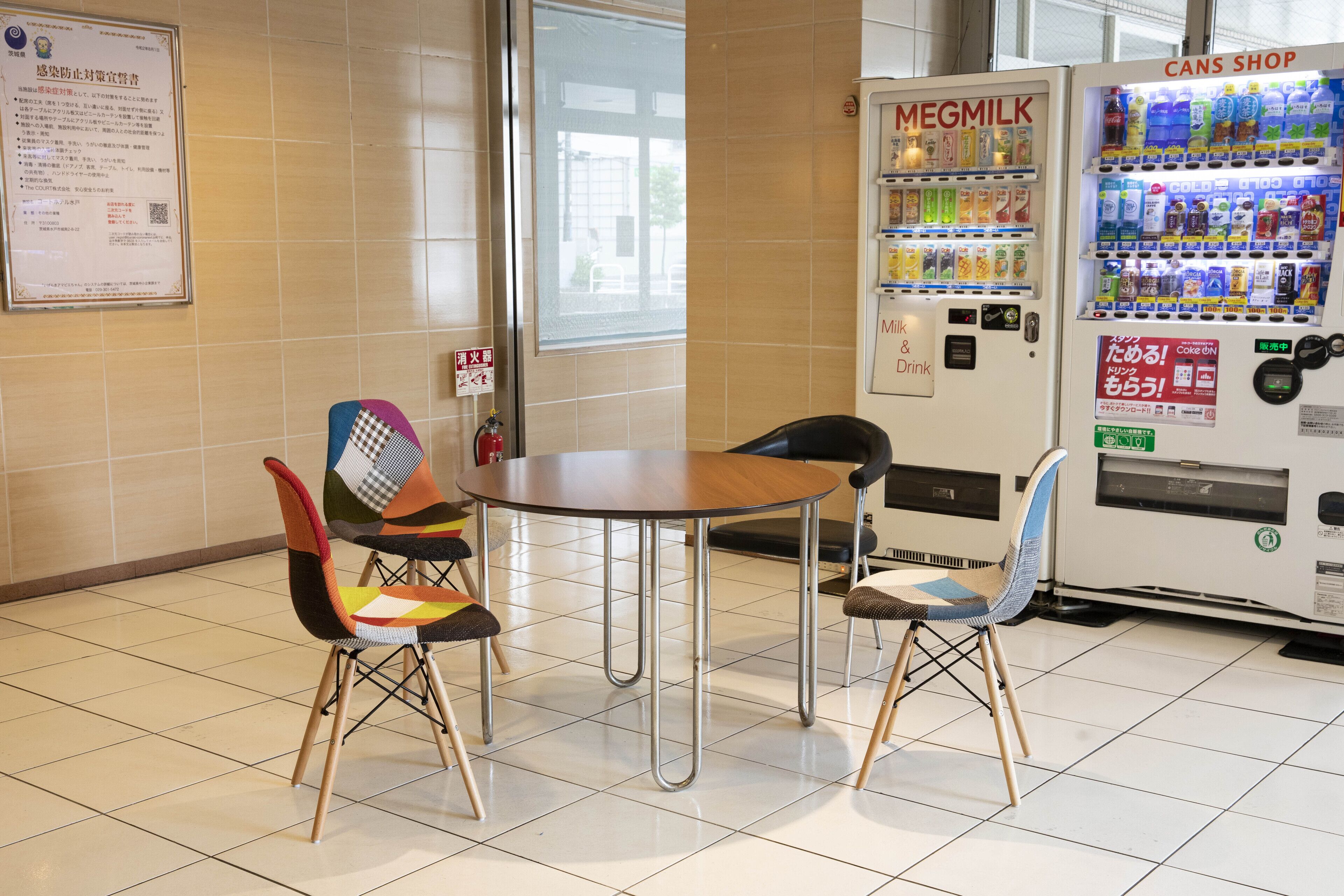 Una cafetería con una mesa redonda y dos sillas de colores, una máquina expendedora de Megmilk y una máquina expendedora de Cans Shop.