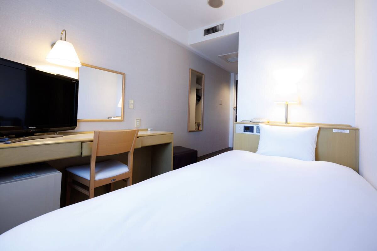 Una habitación de hotel con cama, escritorio, silla, televisor y lámpara de noche.
