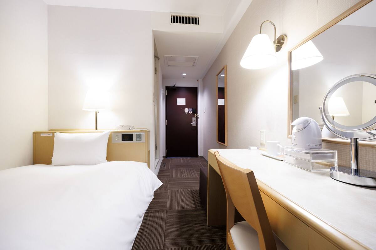 Habitación de hotel con cama, escritorio con silla, espejo redondo y mesita de noche.