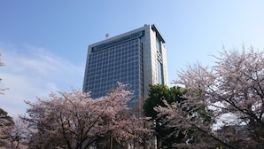 A tall building with a modern glass facade, surrounded by cherry blossom trees. - Court Hotel Mito (Mito)