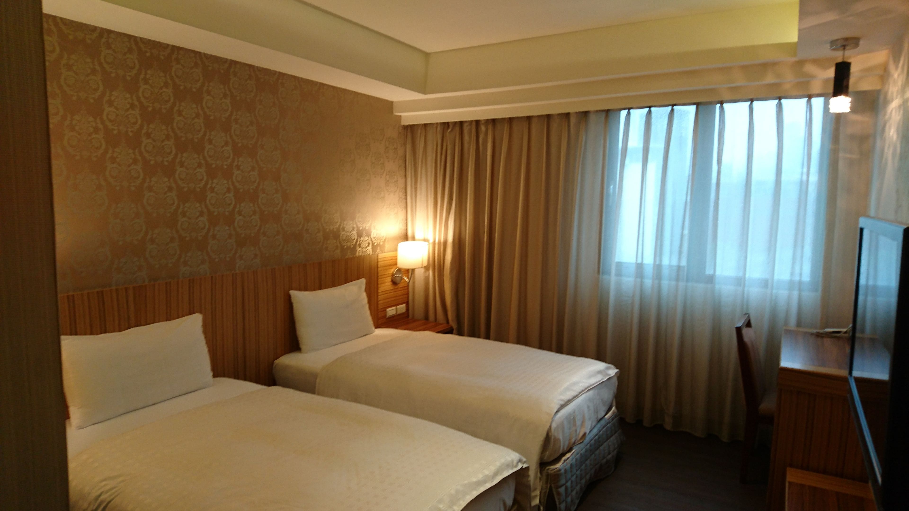 A hotel room with two beds, a desk, a TV, and a window with curtains.