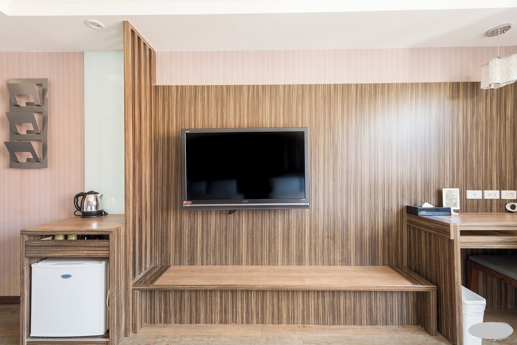 A modern living room with a flat-screen TV mounted on a wooden wall, a small refrigerator, and a coffee maker on a wooden table.