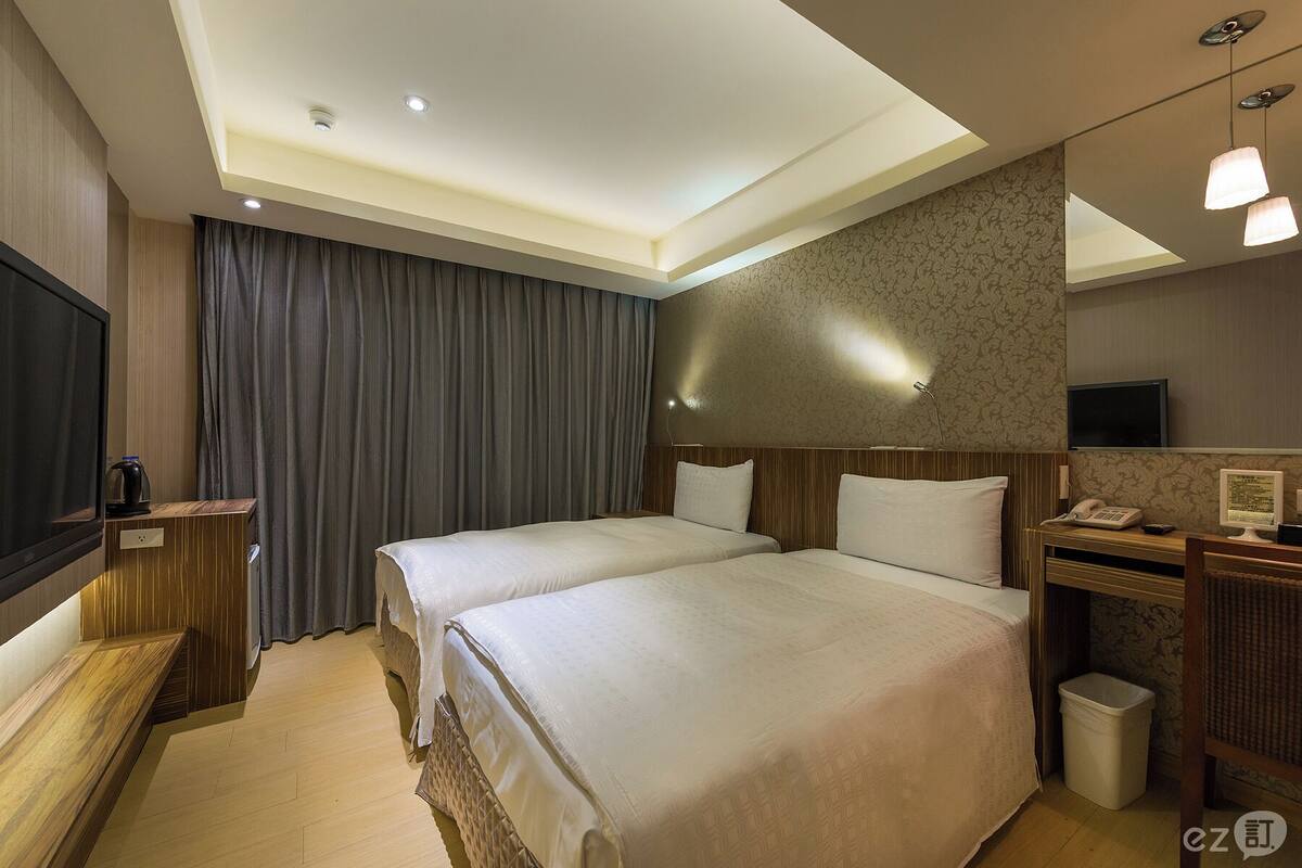 A hotel room with two beds, a desk with a computer, a TV, and a large window with curtains.