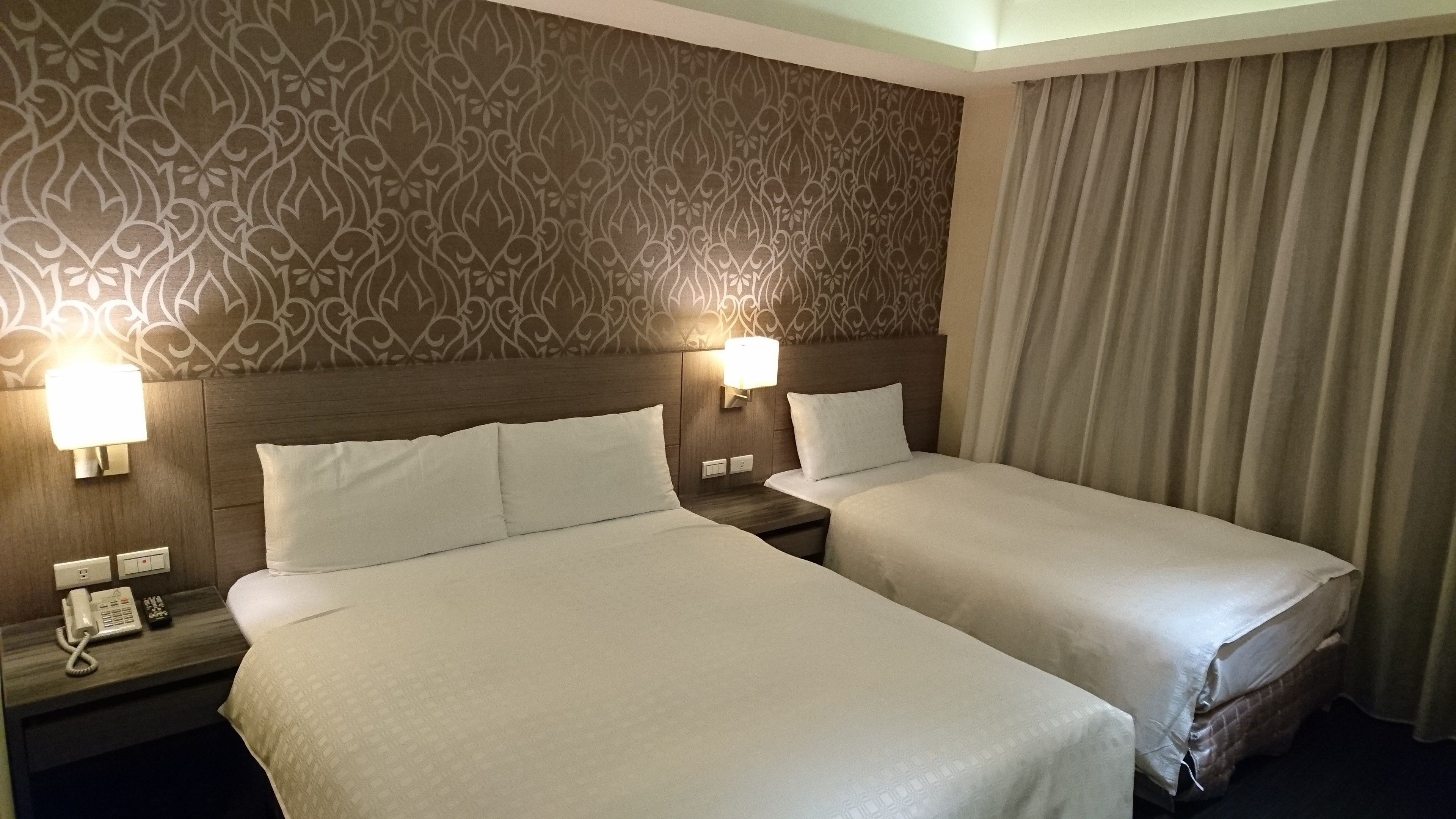 A hotel room with two beds, a patterned wall, a bedside lamp, a phone, and a curtain.