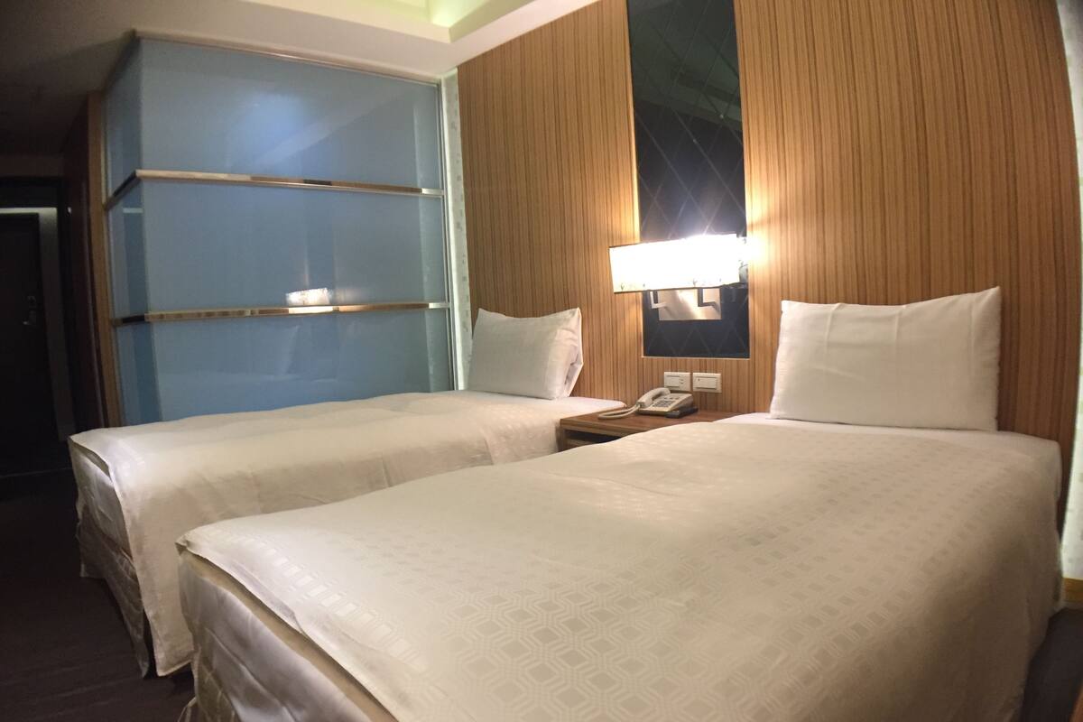 A hotel room with two beds, a nightstand with a phone, and a wall-mounted lamp.