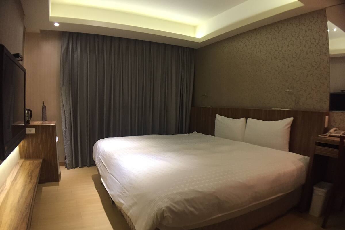 A hotel room with a large bed, a bedside table, a TV, and a curtain.