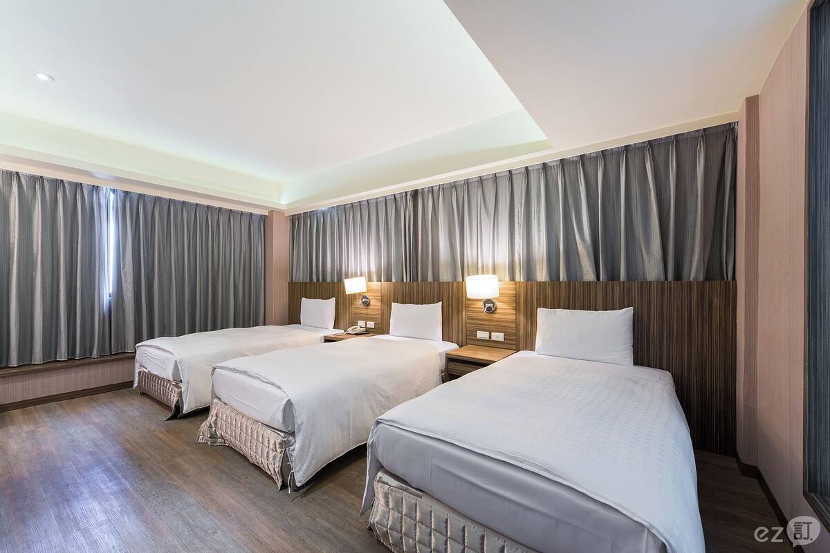 A hotel room with two beds, a wooden headboard, a bedside table with a lamp, and grey curtains.