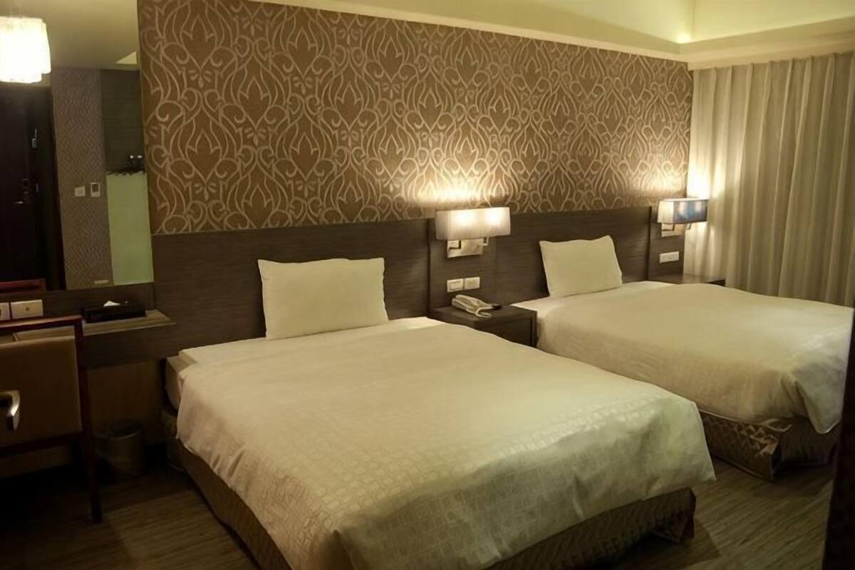 A hotel room with two beds, a nightstand with a lamp, a mirror, and a patterned wall.
