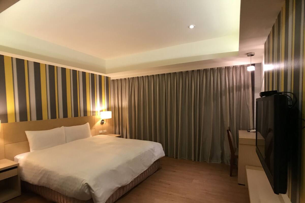 A hotel room with a large bed, a nightstand, a TV, and a curtain.