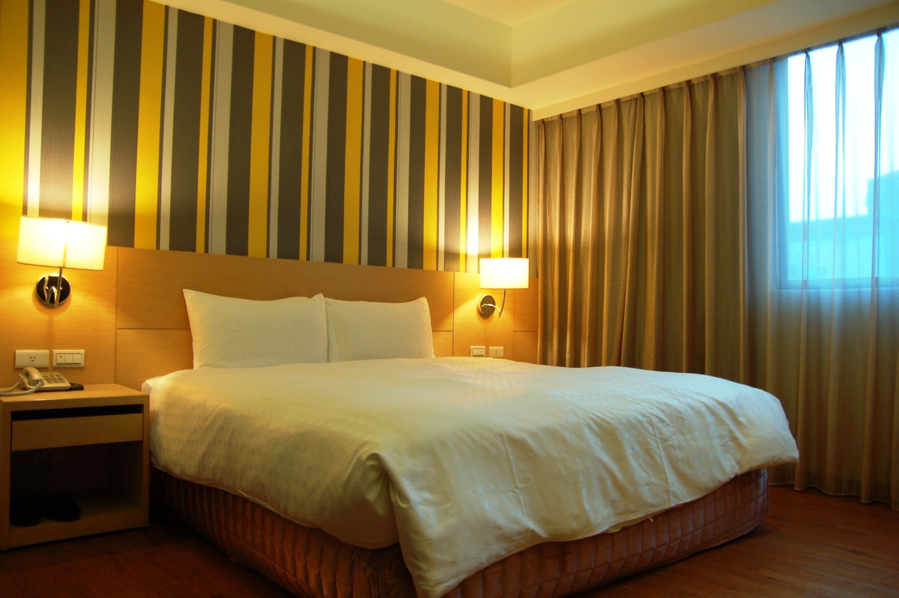 A hotel room with a large bed, two bedside lamps, a nightstand with a phone, and a striped wall.