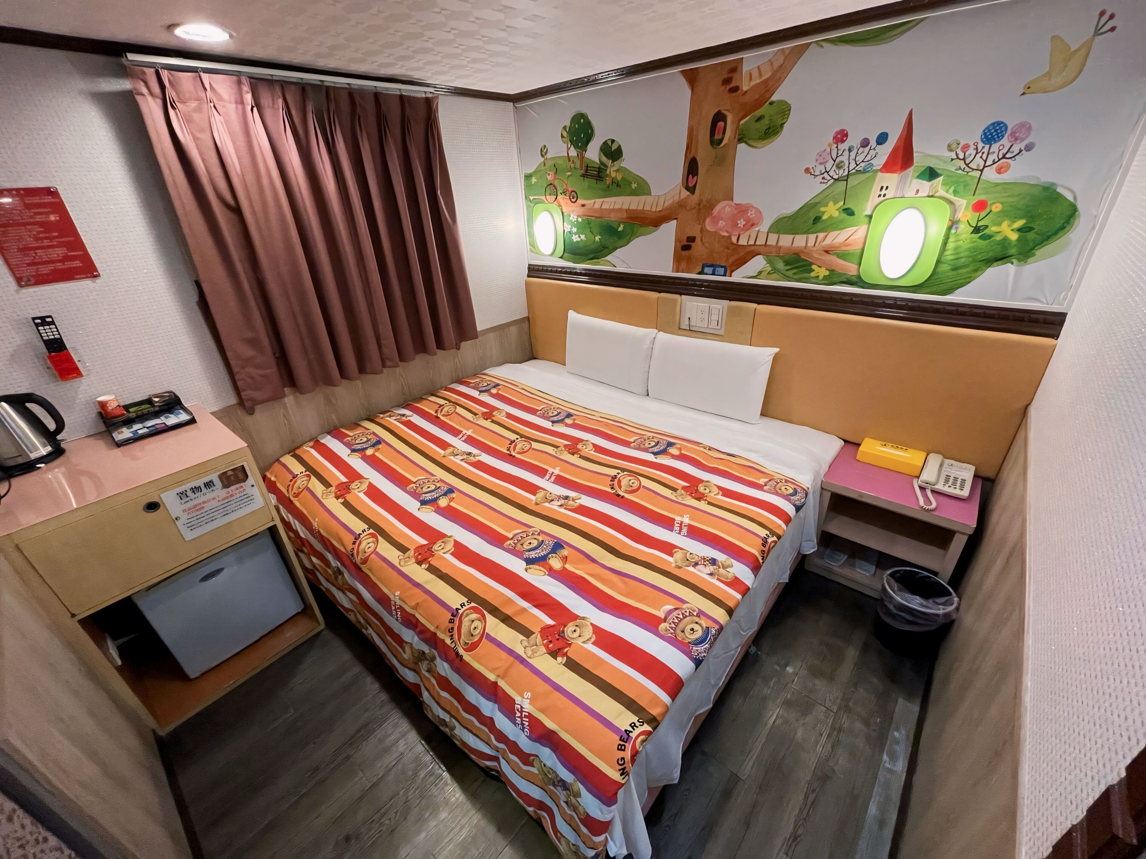 A hotel room with a bed, a desk with a telephone, a trash bin, and a wall with a colorful mural.