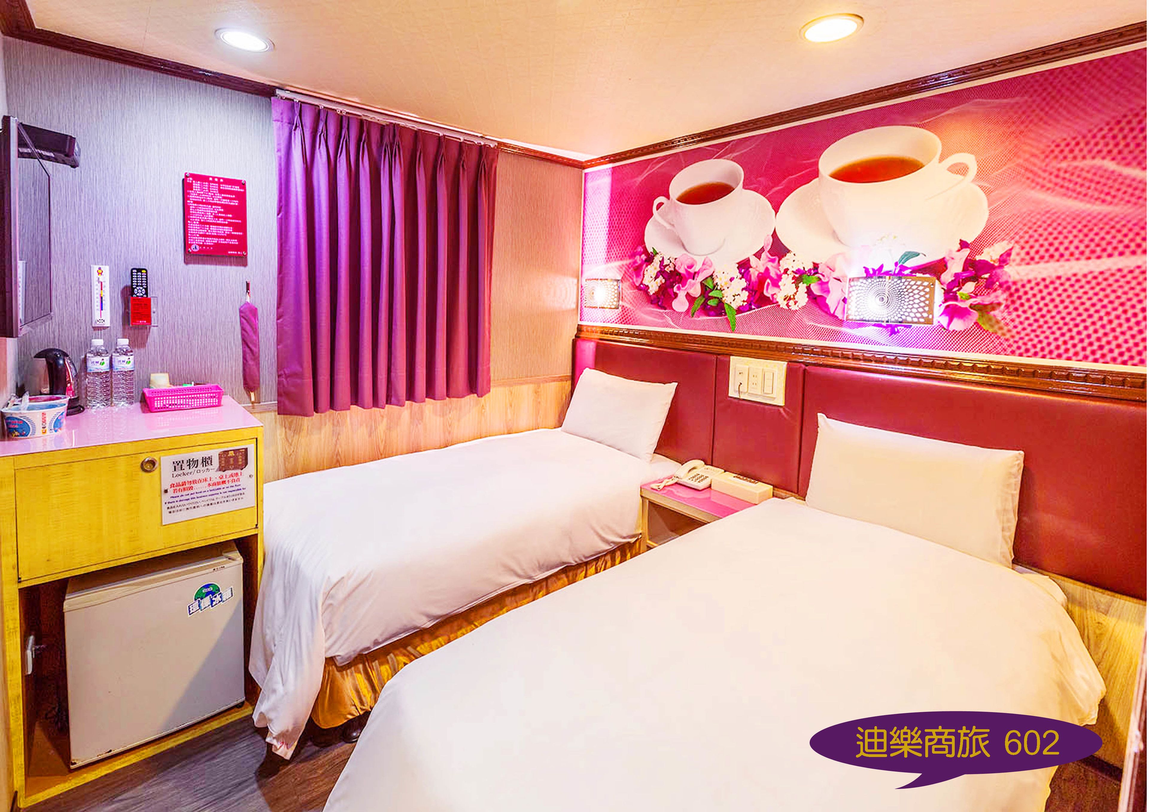 A hotel room with two beds, a desk with a refrigerator, a telephone, and a wall with a floral decoration.
