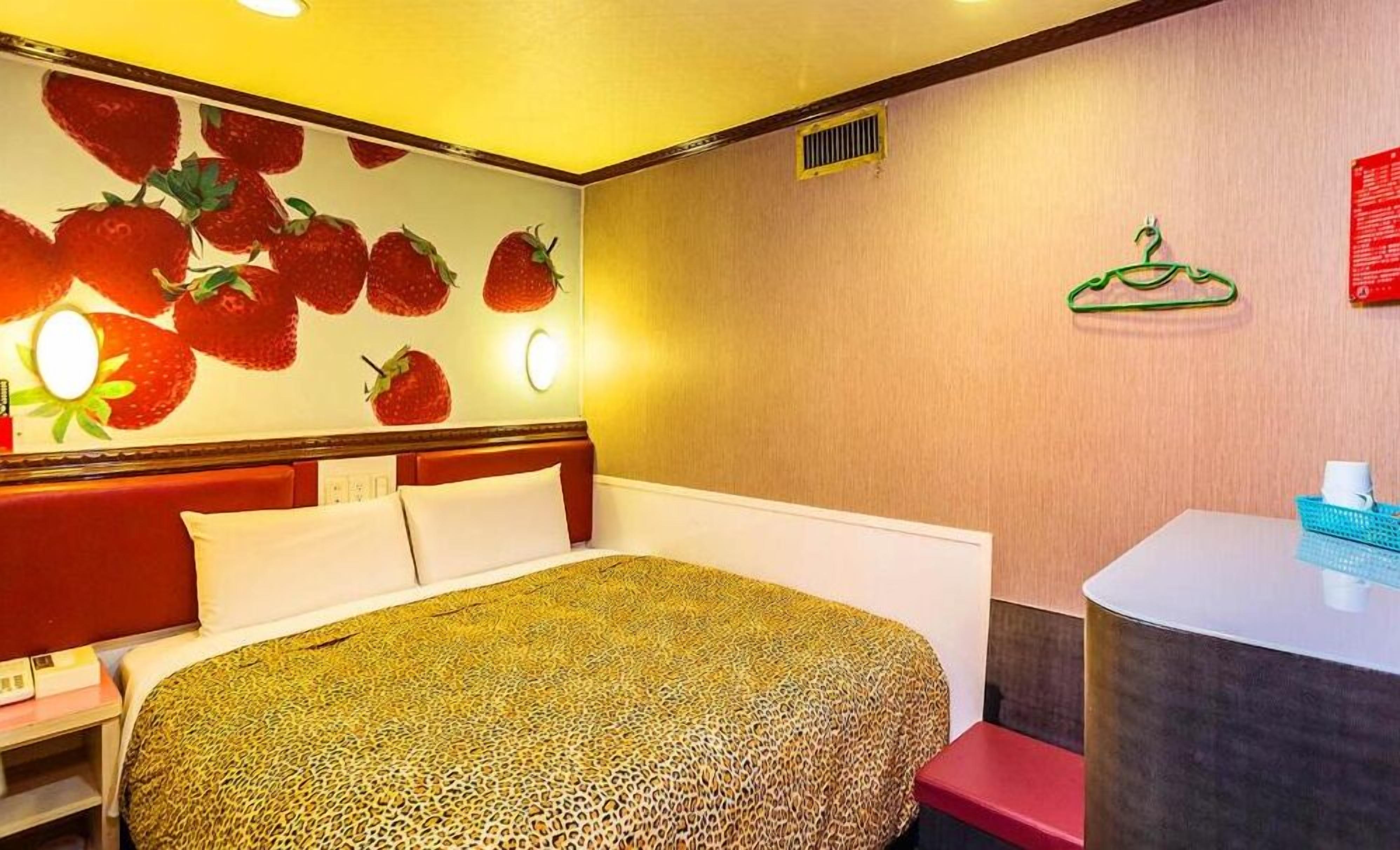A hotel room with a bed, a nightstand, a mirror, a wall with a strawberry mural, a small desk with a chair, and a red bench.