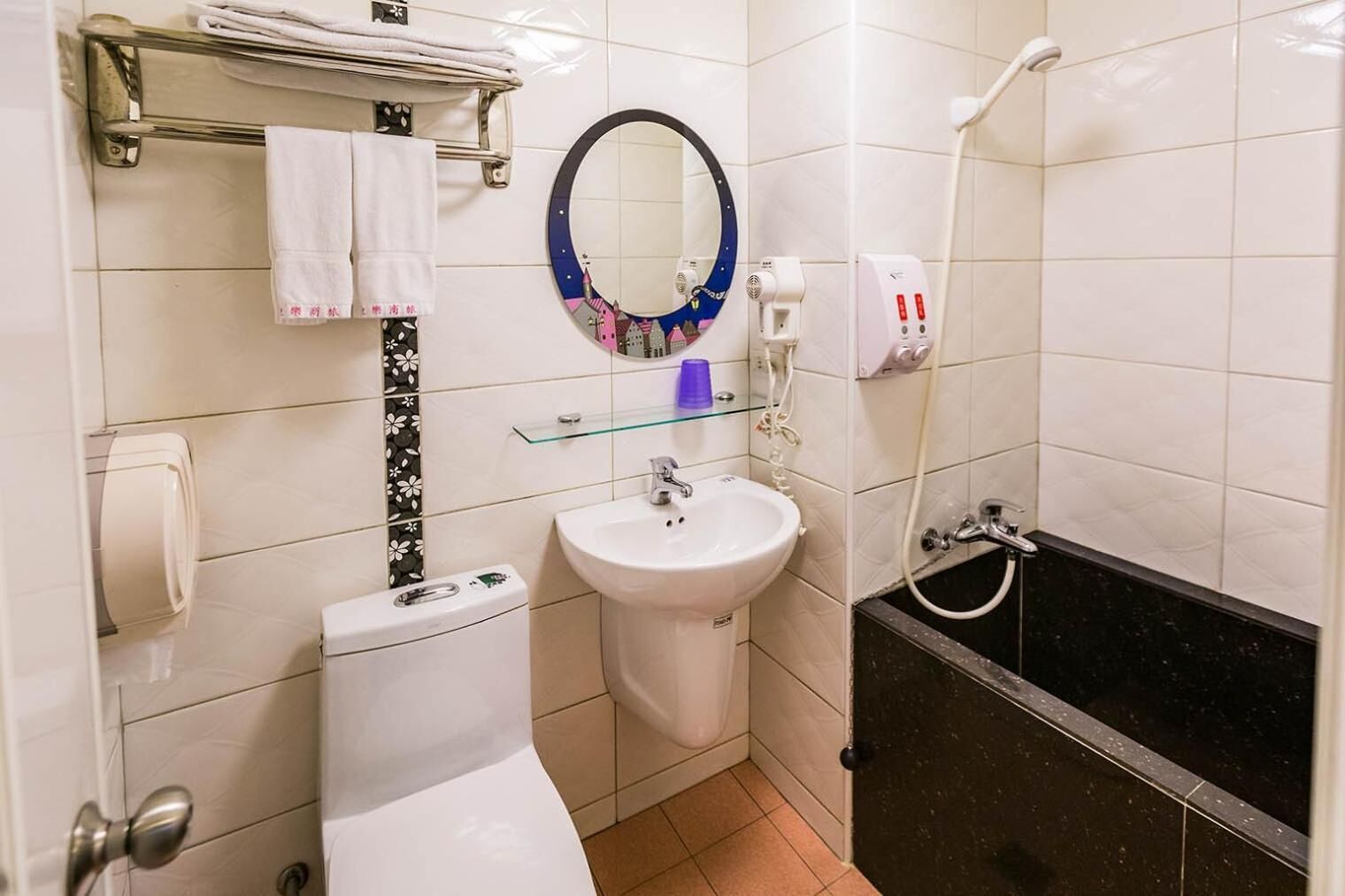 A bathroom with a toilet, sink, and bathtub. There is a towel rack with towels, a glass shelf with toiletries, and a mirror with a decorative frame.