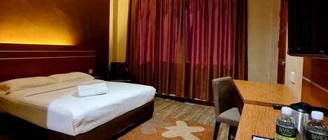 A hotel room with a bed, a desk, a chair, a TV, a window with curtains, and a tray with water bottles and a mug.