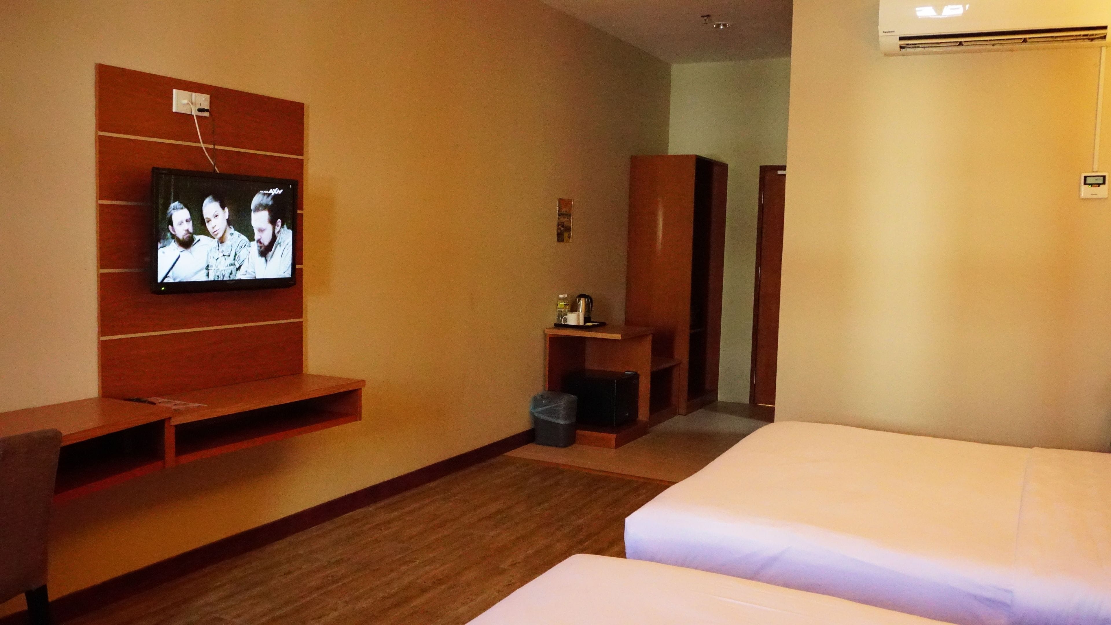 Habitación de hotel con una televisión instalada en la pared, un armario de madera, una mesita con una tetera y una cama.