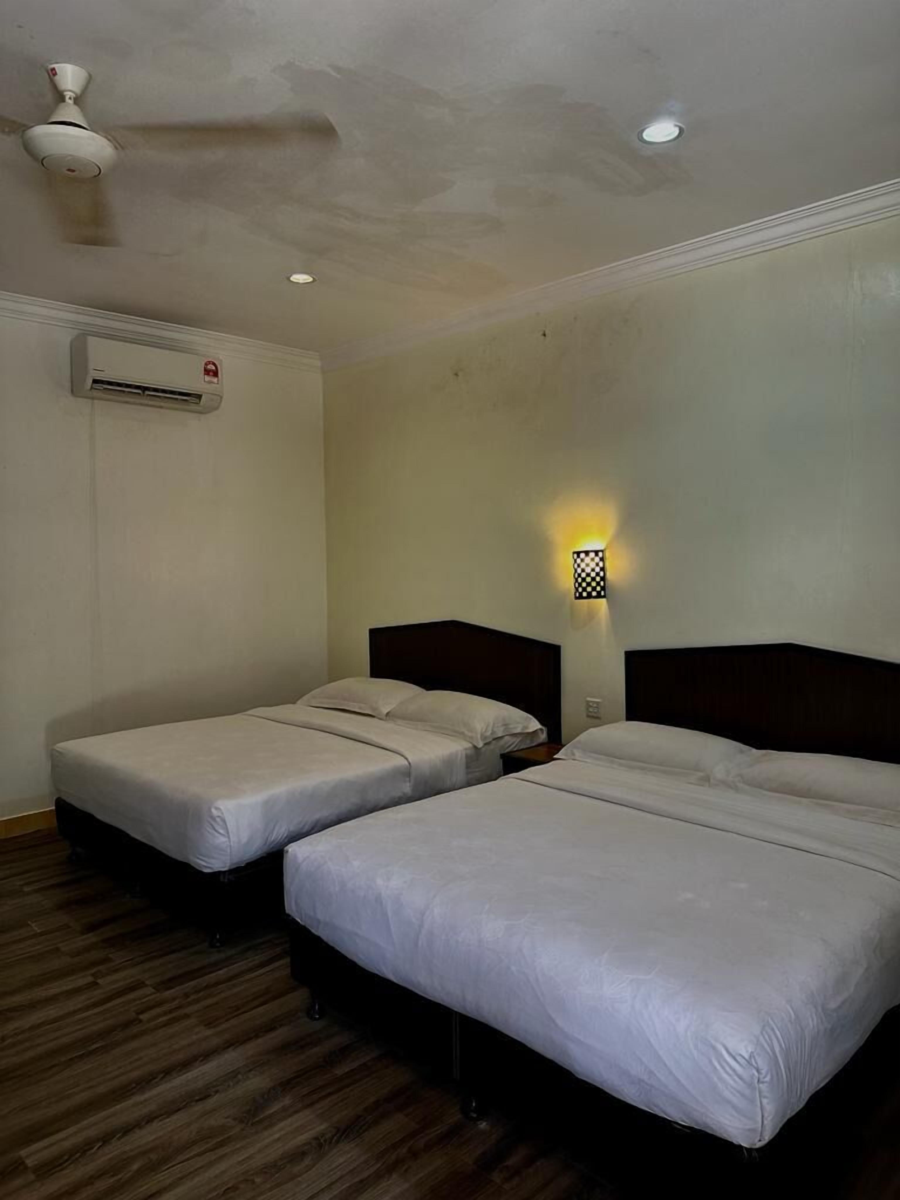 A hotel room with two single beds, a ceiling fan, an air conditioning unit, and a wall-mounted light fixture.