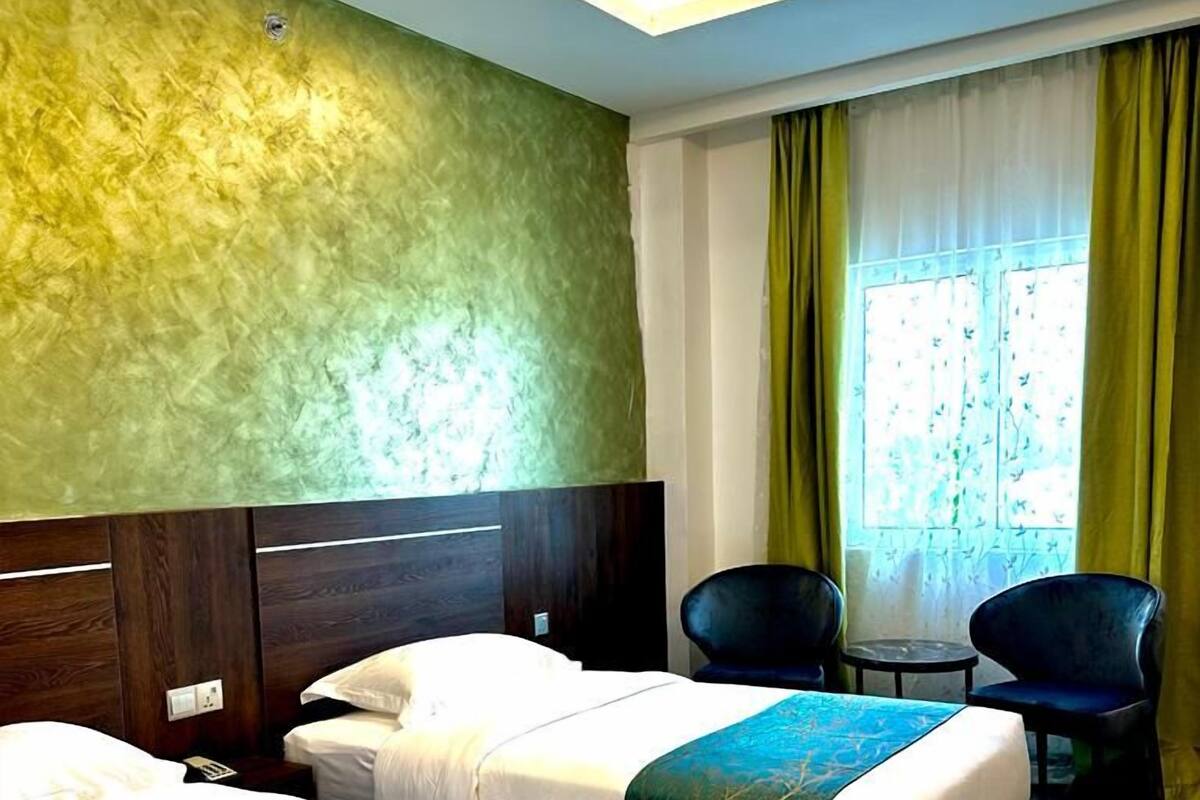 A hotel room with two beds, a wooden floor, a green textured wall, a window with curtains, and a small table with chairs.