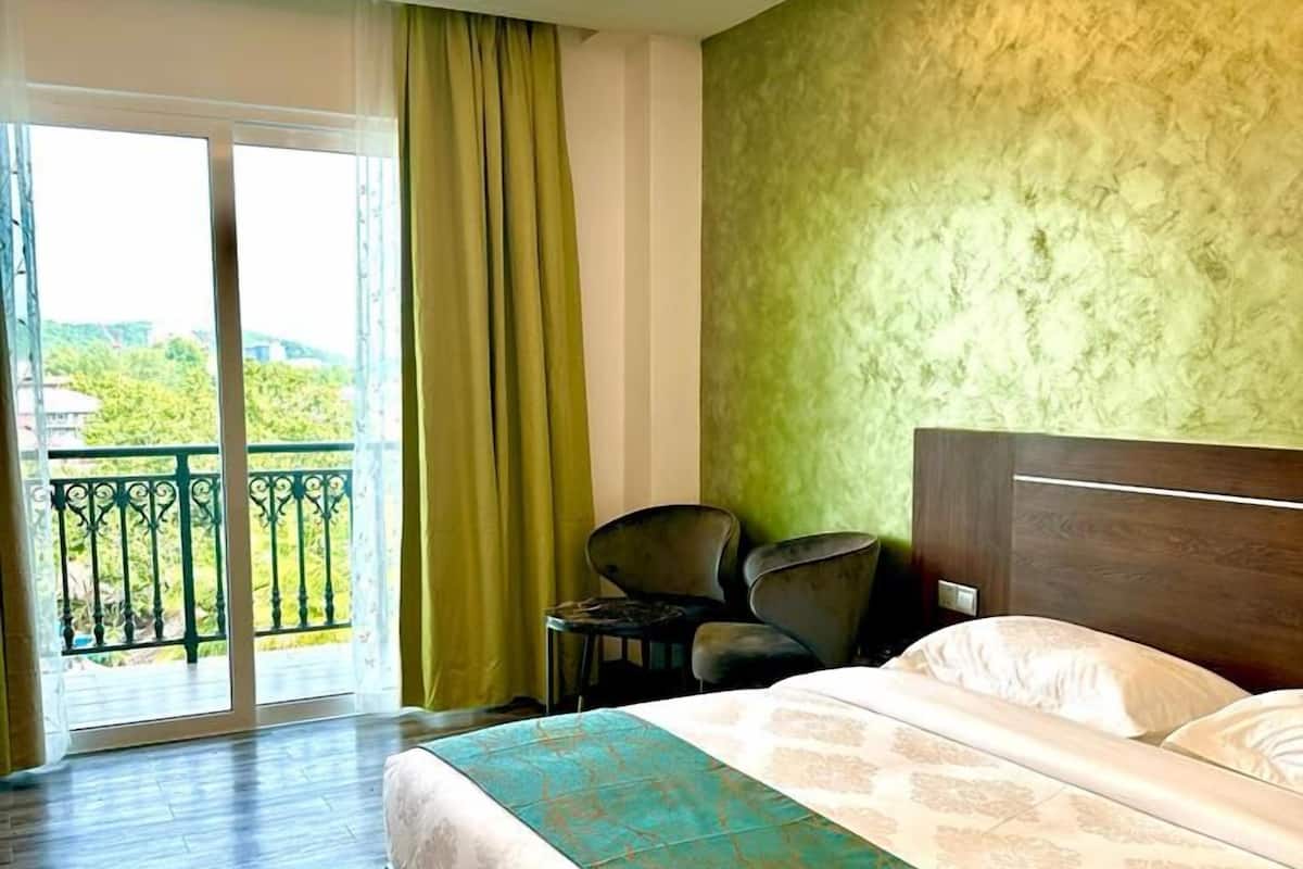 A hotel room with a bed, a chair, a balcony with a view of greenery, and a green textured wall.