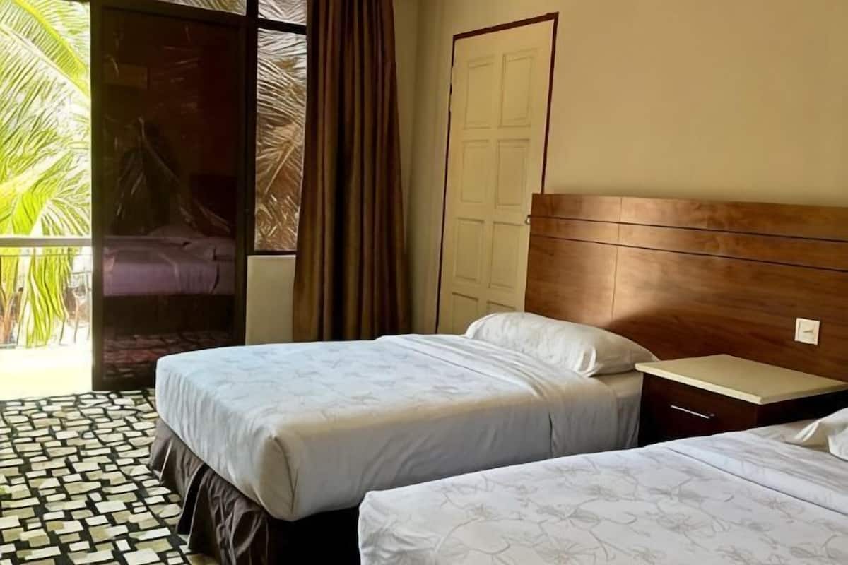 A hotel room with two beds, a wooden headboard, a small bedside table, and a view of a balcony with palm trees.