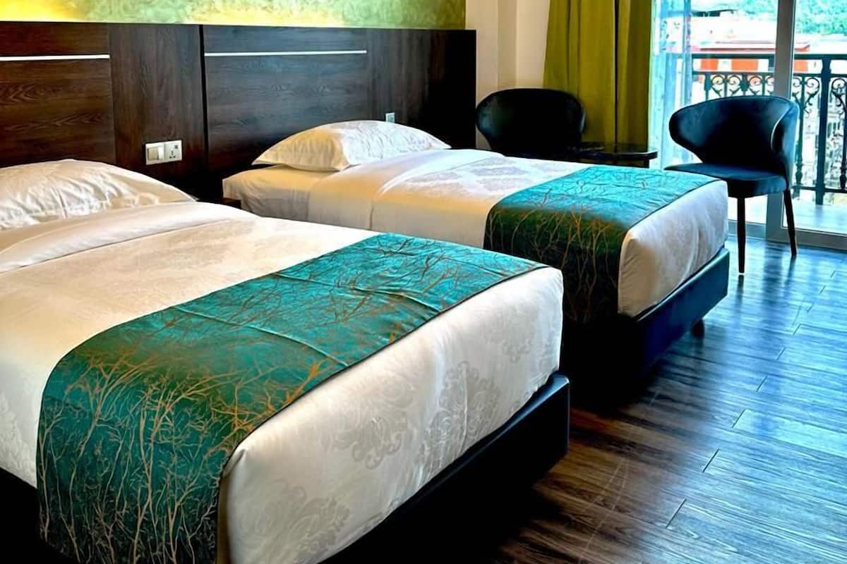 A hotel room with two beds, a wooden floor, a green textured wall, and a view of the outdoors.
