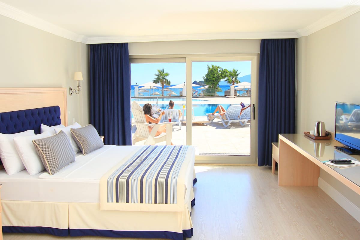 Habitación de hotel con cama, escritorio, televisión y vista a una piscina y playa.