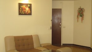 A living room with a brown sofa, a glass coffee table, a door, and a wall-mounted fruit painting. - DMK Donmueang Airport Guesthouse (Pak Kret)
