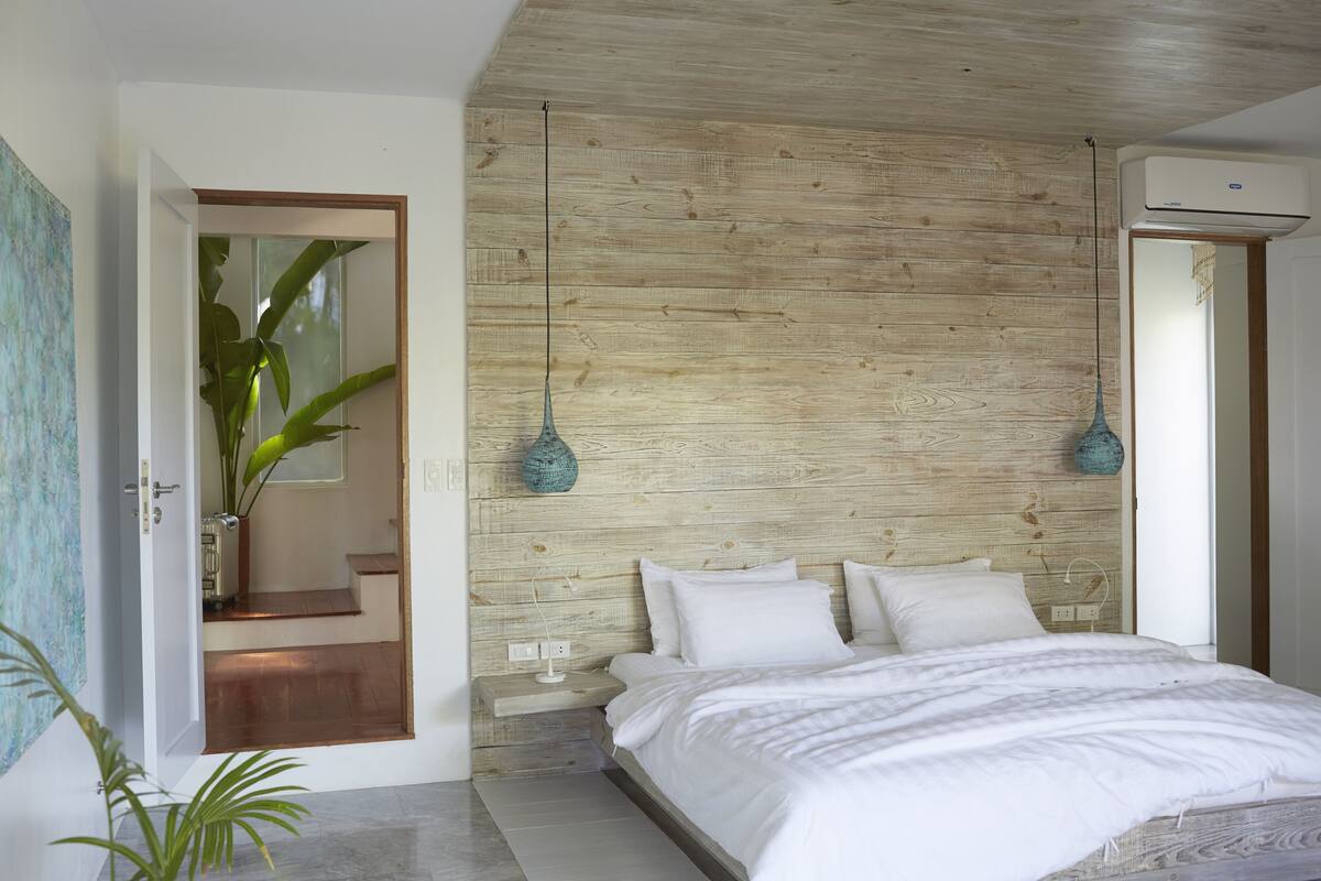 Un dormitorio con cabecera de madera, cama con ropa blanca, lámpara colgante azul y una planta en maceta.