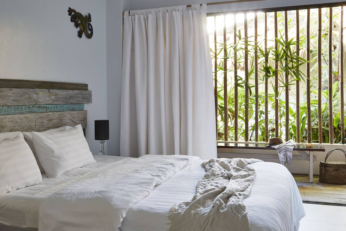 Un dormitorio con cama, cabecera, ventana con cortinas, mesita de noche, lámpara y vistas a plantas de bambú al exterior.